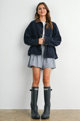 woman wearing a navy corduroy jacket over a white t-shirt and plaid shorts, paired with gray rain boots