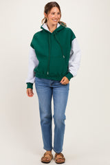 woman wearing a green and gray colorblock maternity hoodie paired with blue jeans and sandals, smiling at the camera