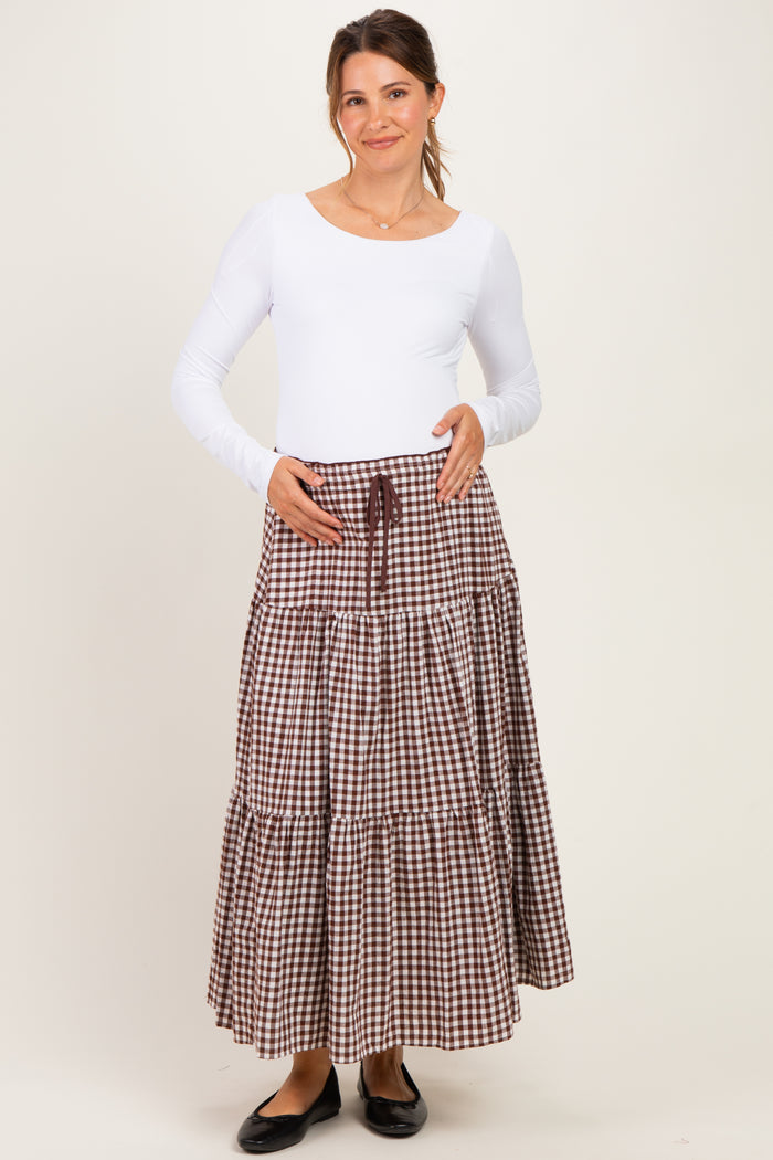 woman wearing a white long sleeve maternity top and a brown gingham tiered maternity skirt, holding her baby bump