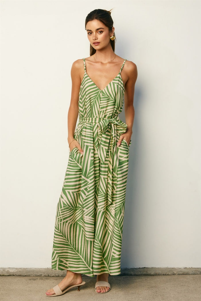 woman wearing a green palm leaf patterned maxi dress with a v-neckline and adjustable tie waist, standing against a wall