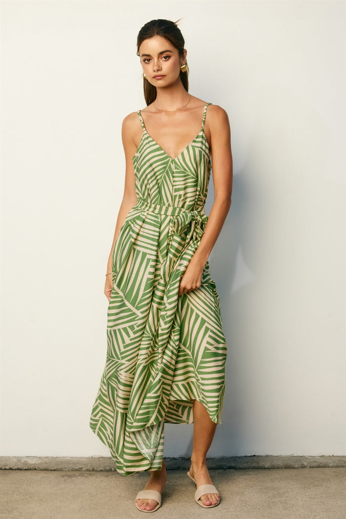 woman wearing a green leaf print maxi dress with spaghetti straps and a gathered waist, standing against a neutral wall