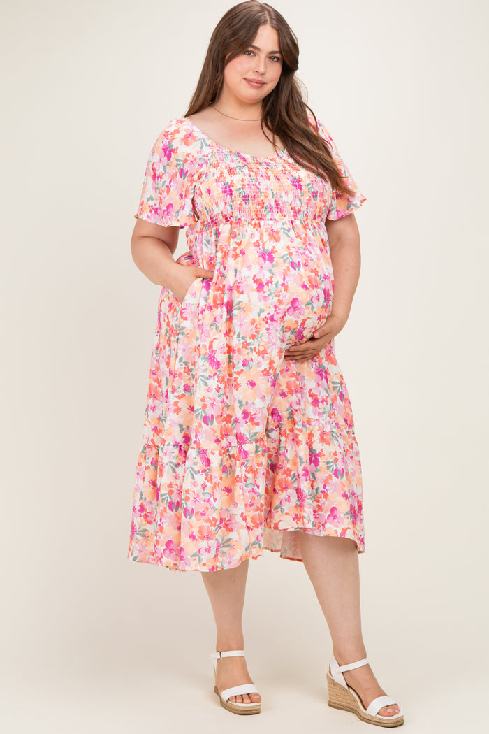 woman wearing a pink floral smocked maternity dress with short sleeves and pockets, holding her baby bump