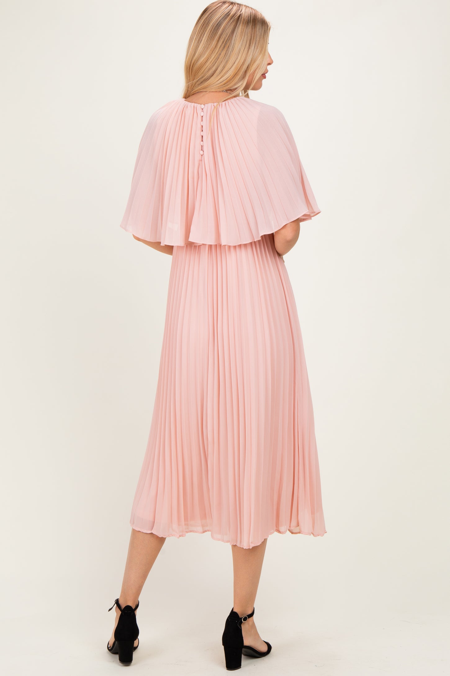 woman wearing a blush pink pleated midi dress with a cape detail and buttons at the back