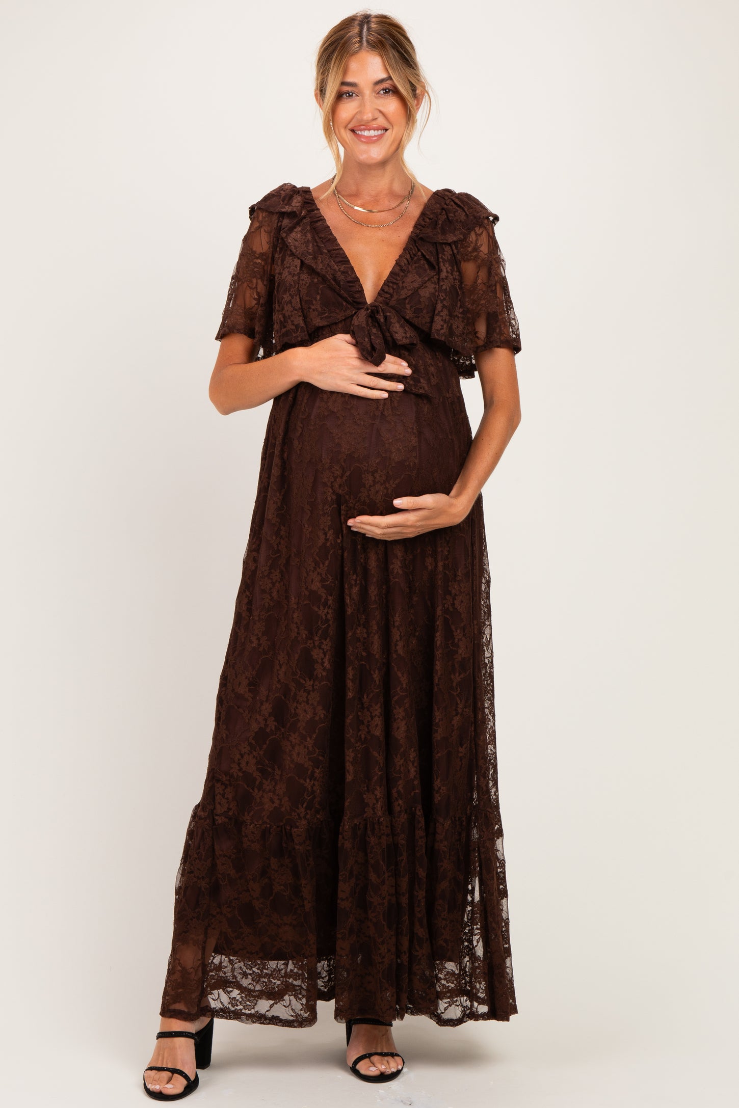 woman wearing a brown lace maternity maxi dress with ruffled sleeves and a bow detail, holding her baby bump