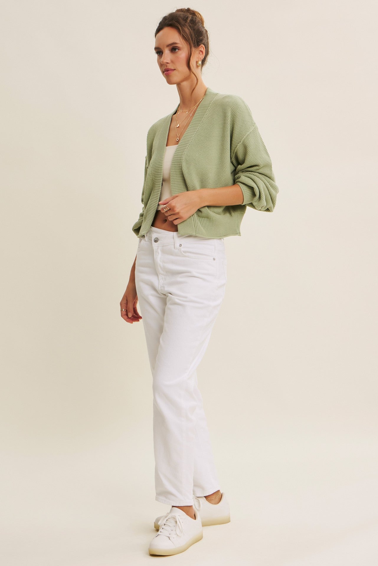 woman wearing a light green cropped cardigan, white jeans, and white sneakers, standing against a neutral background