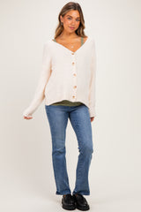 woman wearing a cream maternity cardigan with buttons over a green tank top and blue jeans, smiling and standing casually