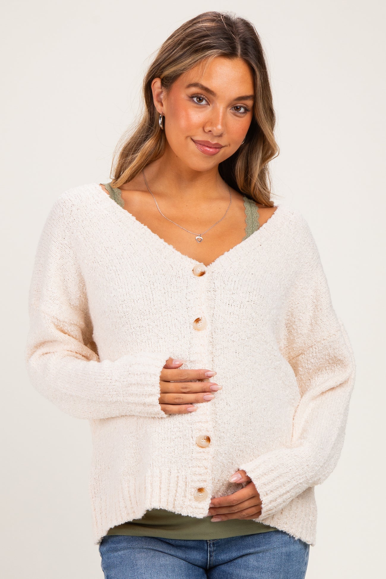 woman wearing a cream maternity cardigan with buttons, holding her baby bump, styled over a green tank top and jeans