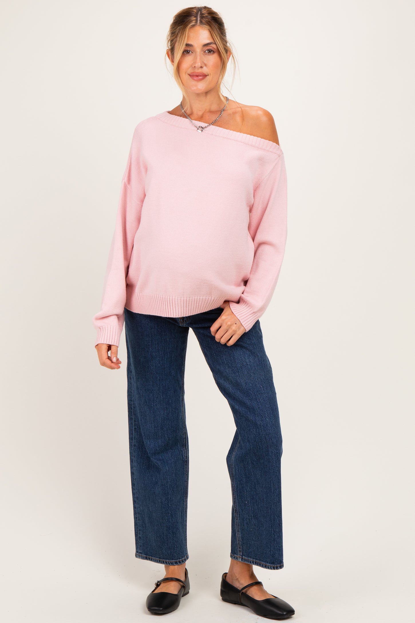 woman wearing a pink off-shoulder maternity sweater with a baby bump and blue wide-leg jeans