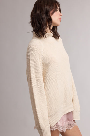 woman wearing a cream oversized knit sweater with a high neckline and relaxed fit over a light pink lace trim top