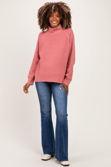 woman wearing a pink maternity sweater with a high neckline and flared jeans, smiling and showing her baby bump