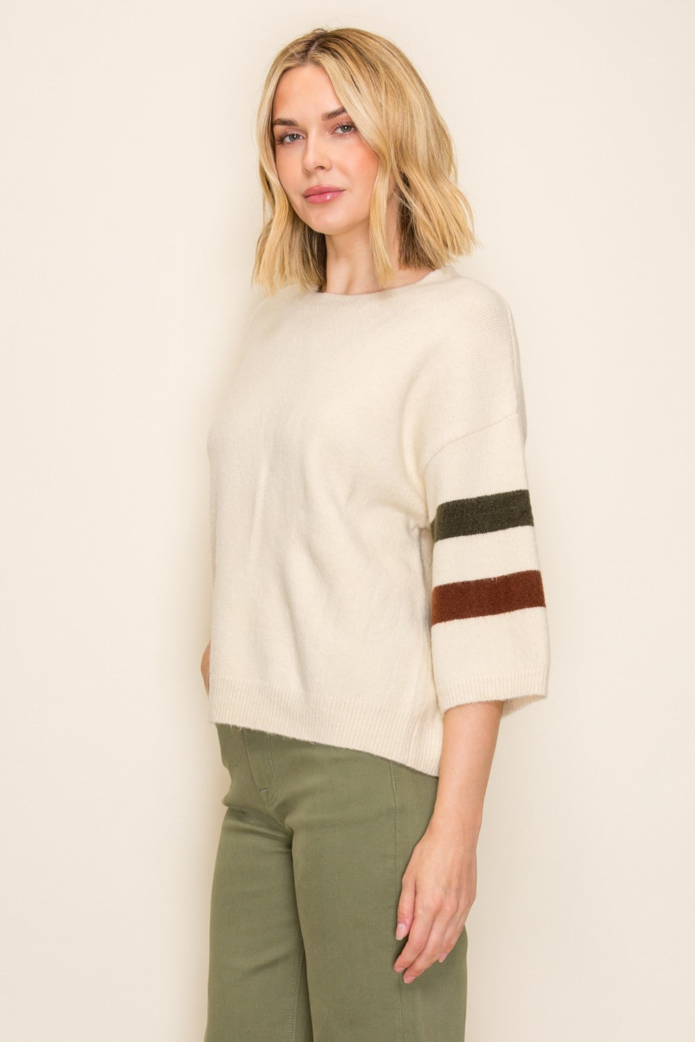 woman wearing an ivory oversized sweater with color block stripes on the sleeves and green trousers