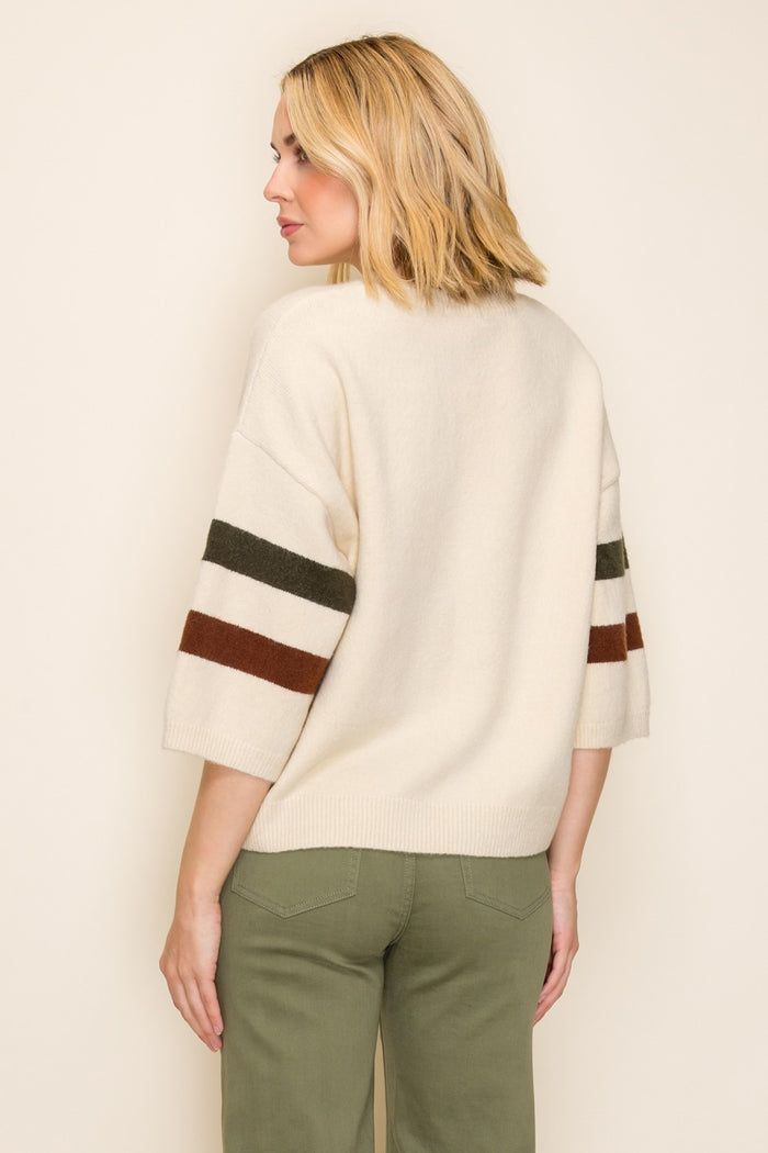 woman wearing a cream oversized sweater with brown and green stripes on the sleeves, styled with olive green pants
