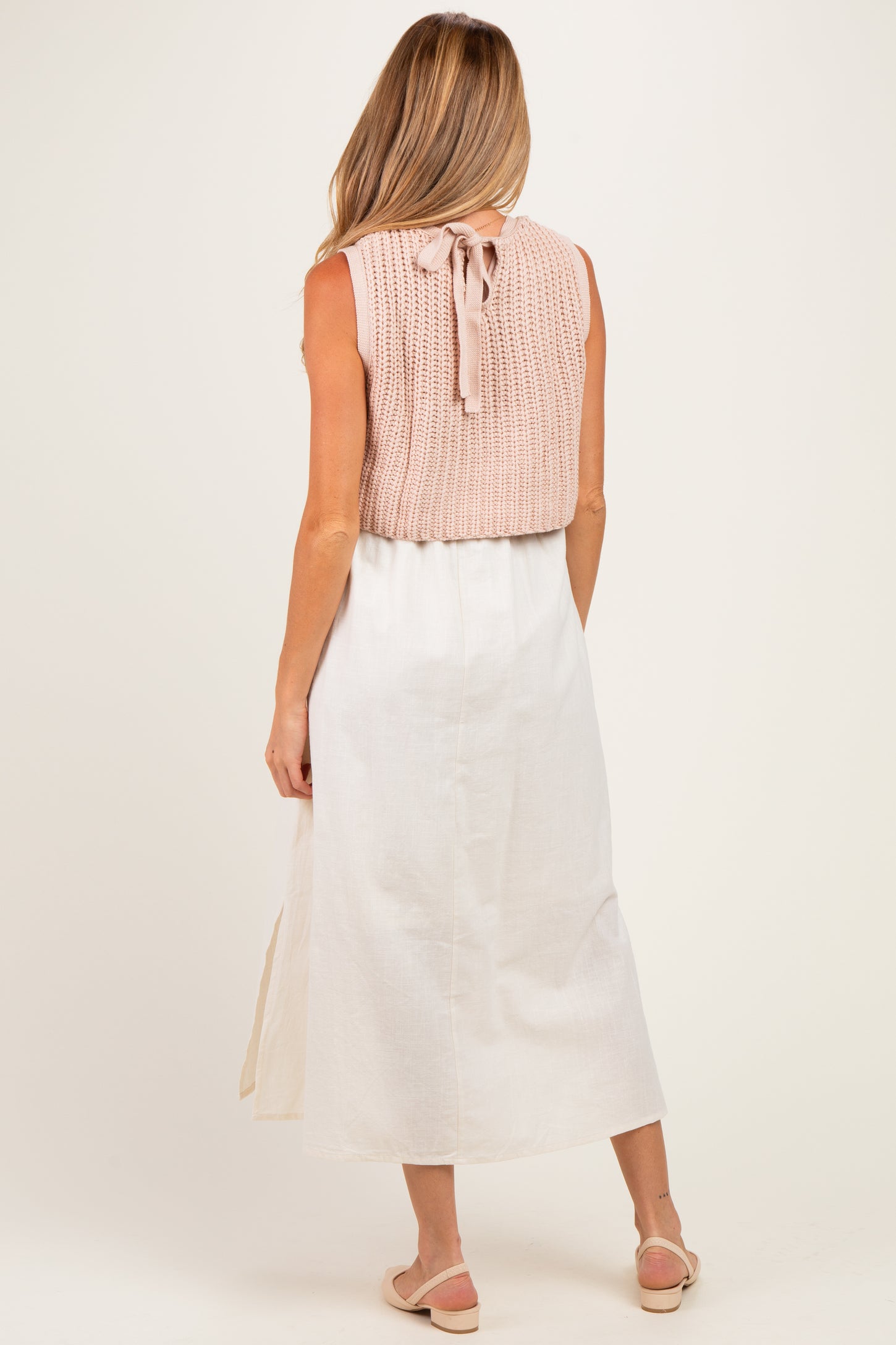 woman wearing a light pink knitted maternity top with a tie at the back and a white maxi skirt, standing with her back to the camera