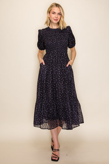 woman wearing a black floral midi dress with puff sleeves and pockets, standing against a neutral backdrop