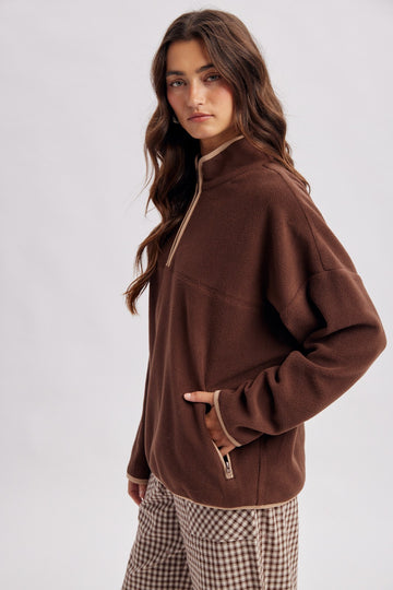 woman wearing a brown zip-up fleece pullover with a high neck and contrasting trim, paired with checked pants