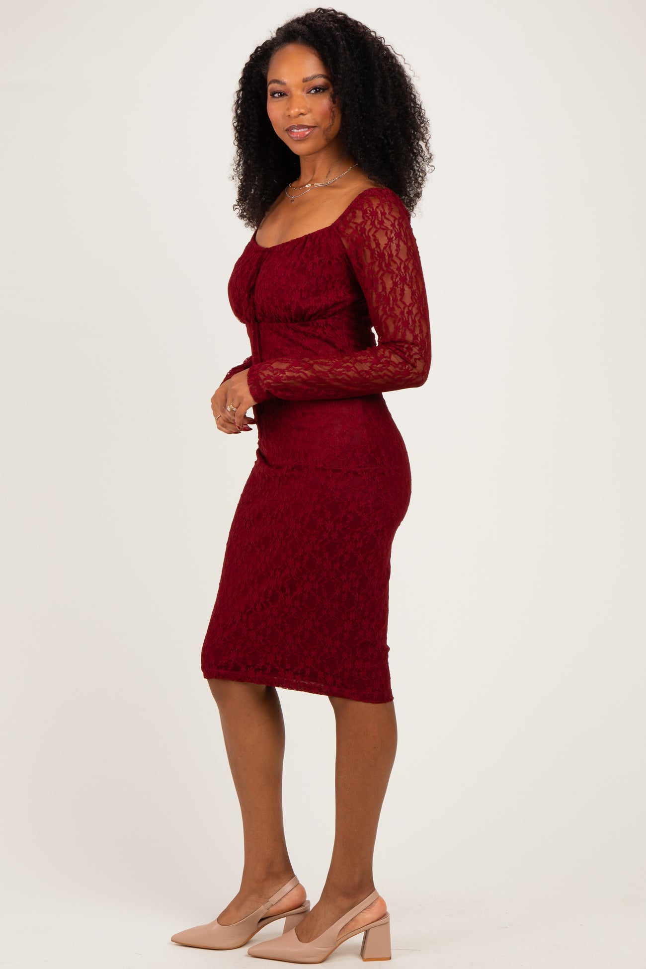 woman wearing a burgundy lace long sleeve bodycon dress and nude heels, standing sideways with a slight smile