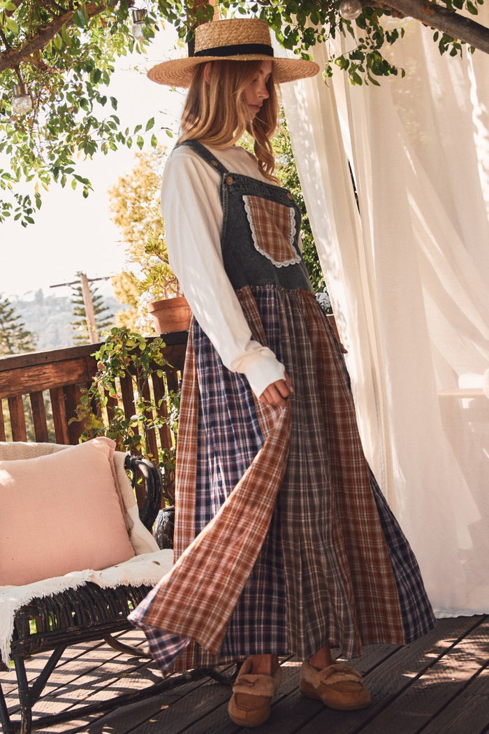 woman wearing a denim overalls dress with a plaid skirt and a white long sleeve top, standing outdoors in natural light