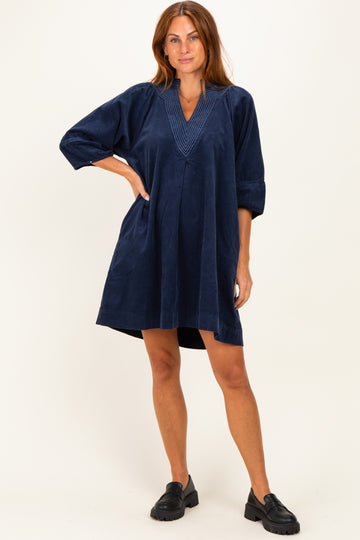 woman wearing a navy blue short sleeve dress with a v-neck and pleated detailing, standing with one hand on her hip