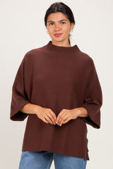 woman wearing a brown oversized sweater with a high neckline and wide sleeves, standing against a light background
