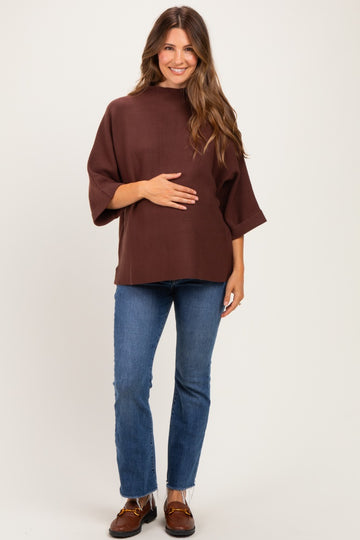 woman wearing a brown maternity top with three-quarter sleeves and holding her baby bump, paired with blue skinny jeans