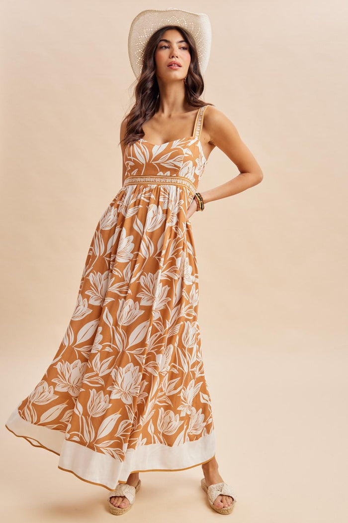 woman wearing an orange floral maxi dress with a square neckline and straps, posing stylishly with a wide-brimmed hat