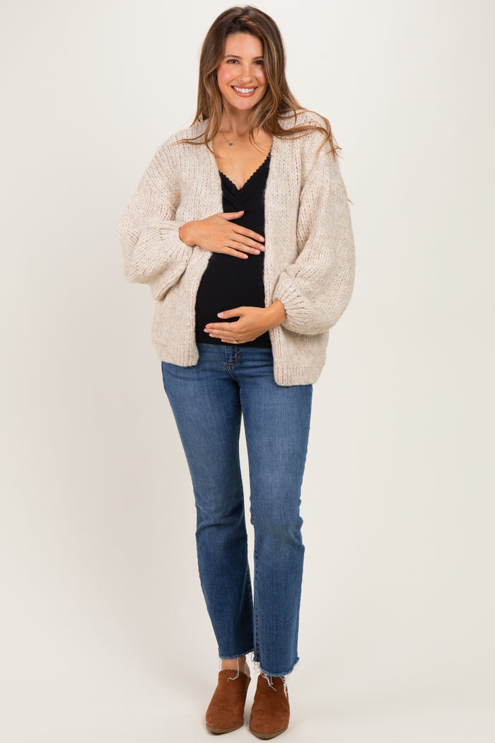 woman wearing a beige knitted maternity cardigan and black top, holding her baby bump with a smile