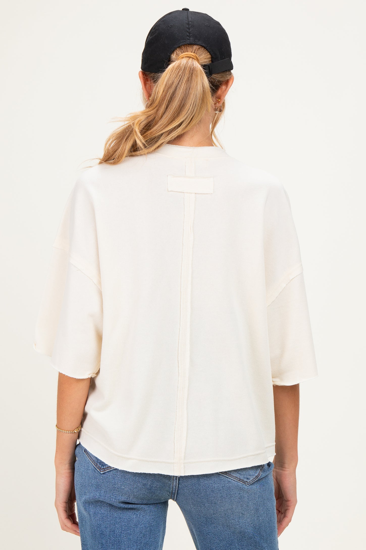 woman wearing a cream-colored maternity top with a relaxed fit and back seam detail, styled with a black cap