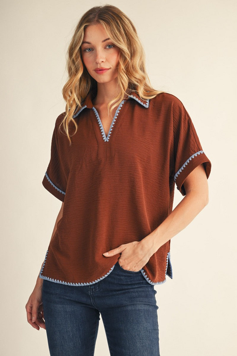 woman wearing a brown short sleeve top with blue trim and a v-neckline, posing with one hand on her hip