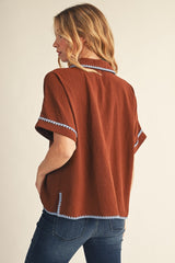 woman wearing a brown short sleeve top with contrasting blue embroidery details, styled with denim jeans