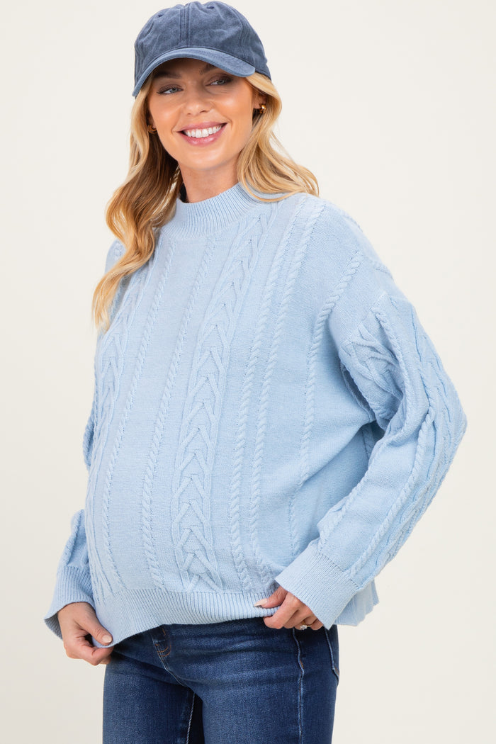 woman wearing a light blue cable knit maternity sweater and a navy cap, showcasing her baby bump