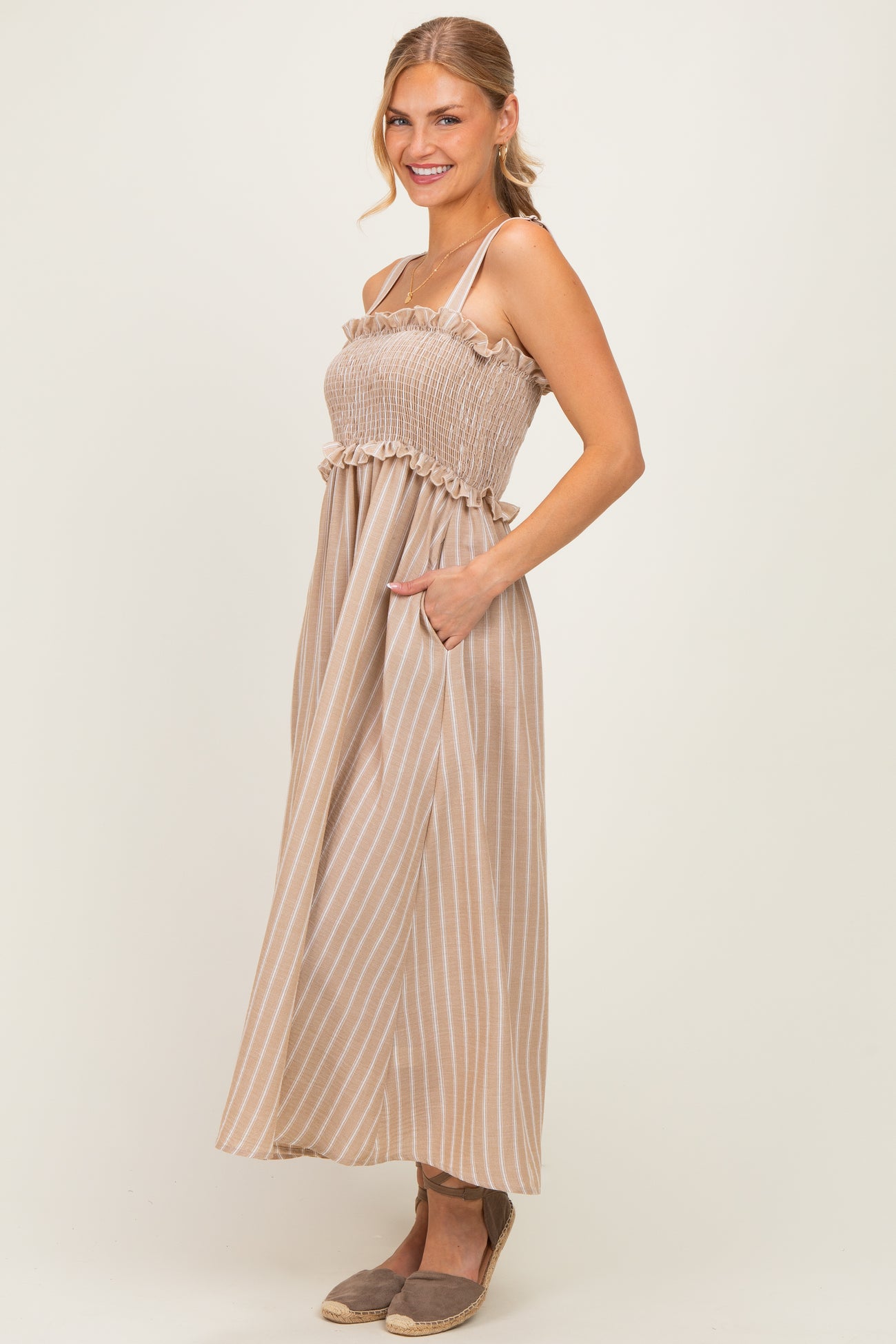 woman wearing a beige and white striped maxi dress with a smocked bodice and ruffled straps, posing with hands in pockets