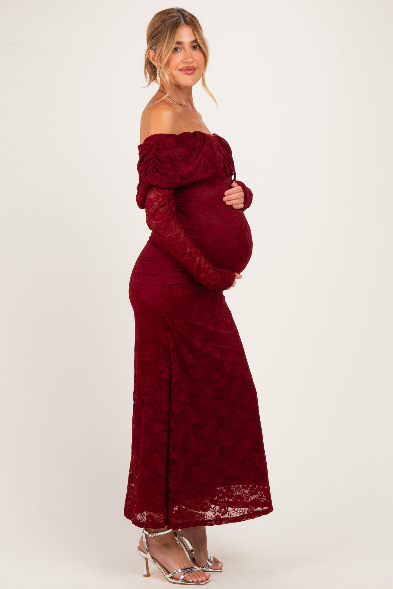 woman wearing a burgundy off-shoulder lace maternity dress with long sleeves and holding her baby bump