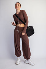 woman wearing a brown long sleeve crop top and matching wide-leg pants, holding a phone and wearing sneakers