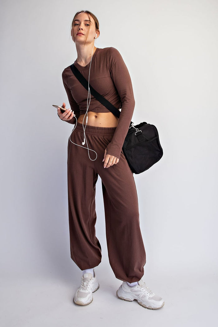 woman wearing a brown long sleeve crop top and matching wide-leg pants, holding a phone and wearing sneakers
