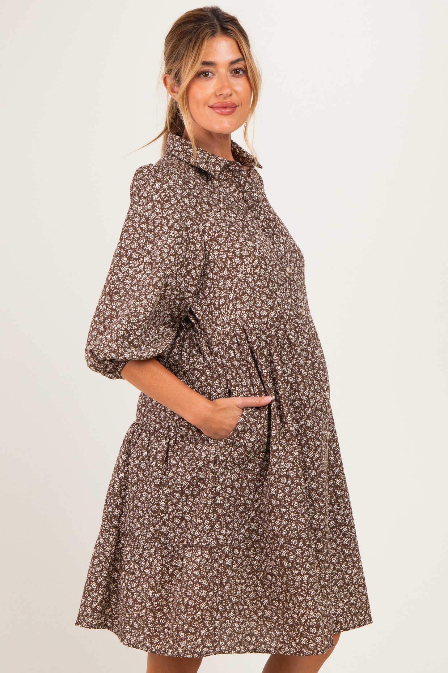 woman wearing a brown floral maternity dress with puff sleeves and side pockets, posing with a visible baby bump