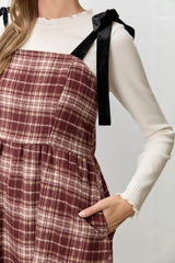 woman wearing a burgundy plaid pinafore dress with black velvet straps over a cream ribbed long sleeve top