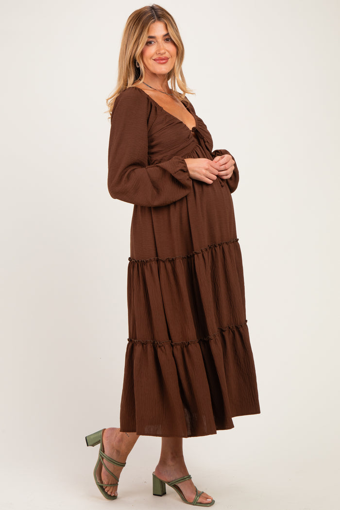 woman wearing a brown maternity maxi dress with long sleeves and tiered ruffles, holding her baby bump