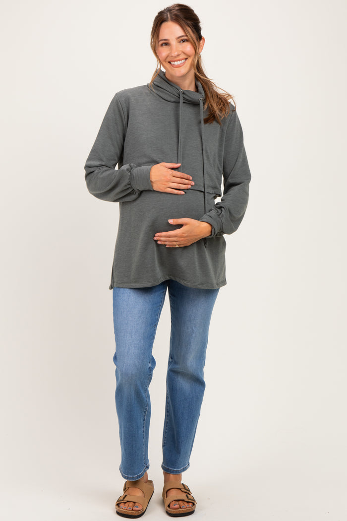 woman wearing a dark gray maternity hoodie with a cowl neck and holding her baby bump while standing in casual blue jeans