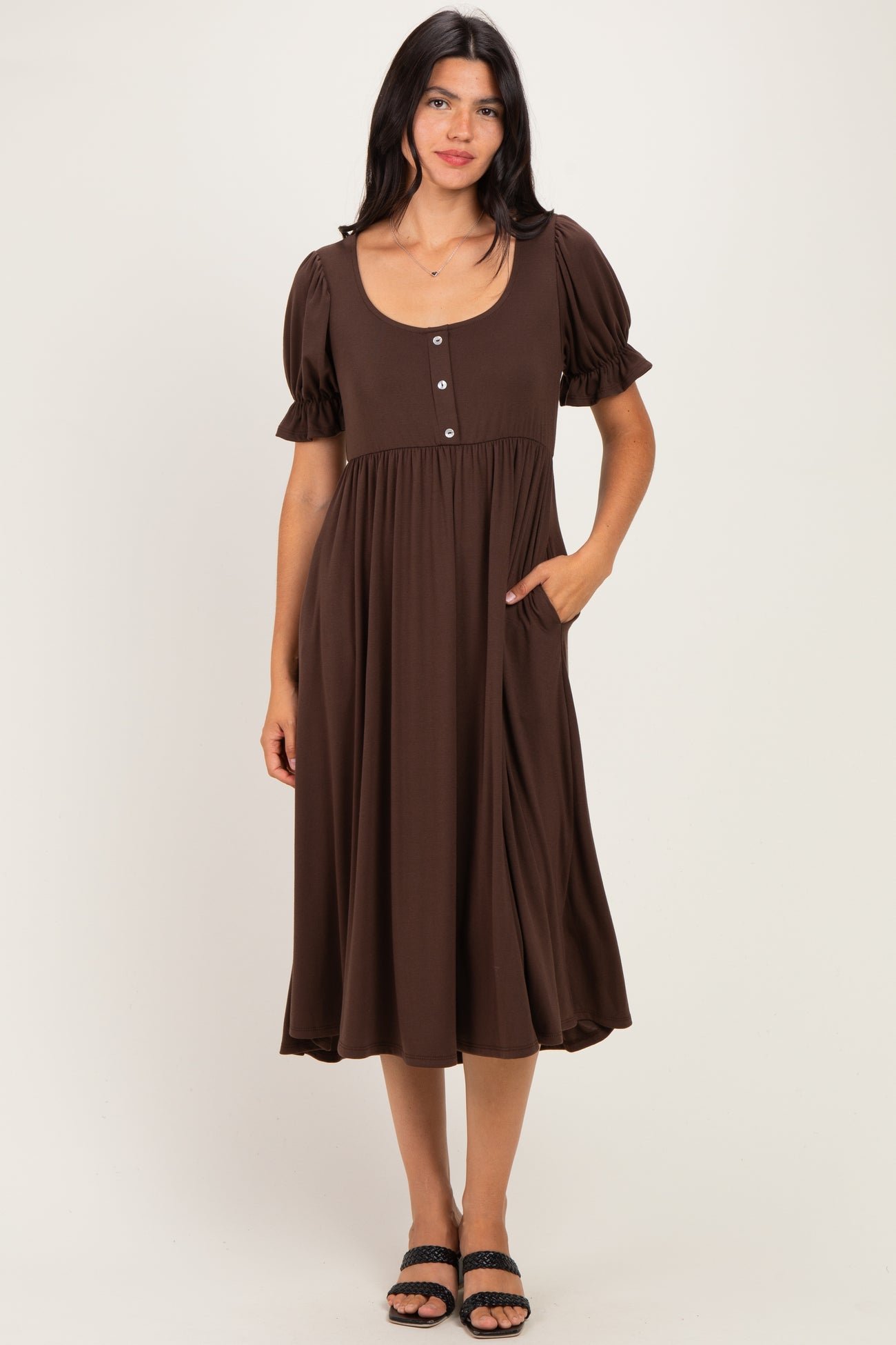 woman wearing a brown midi dress with puff sleeves, a scoop neckline, and button details, standing with one hand in her pocket