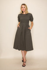 woman wearing a dark gray midi dress with puff sleeves and pockets, standing against a neutral background