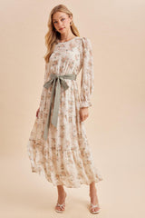 woman wearing a floral maxi dress with long sleeves and a tie waist in soft pastel colors, posing against a neutral background