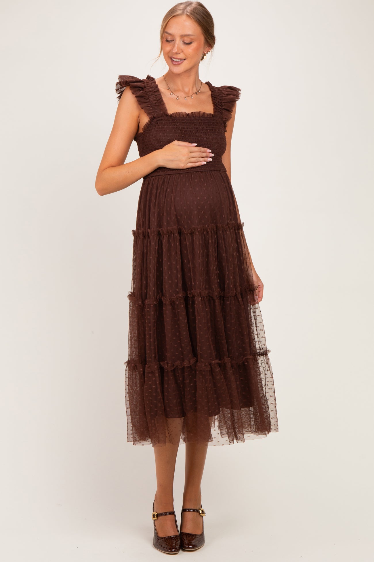 woman wearing a brown maternity dress with ruffled straps and tiered tulle, holding her baby bump