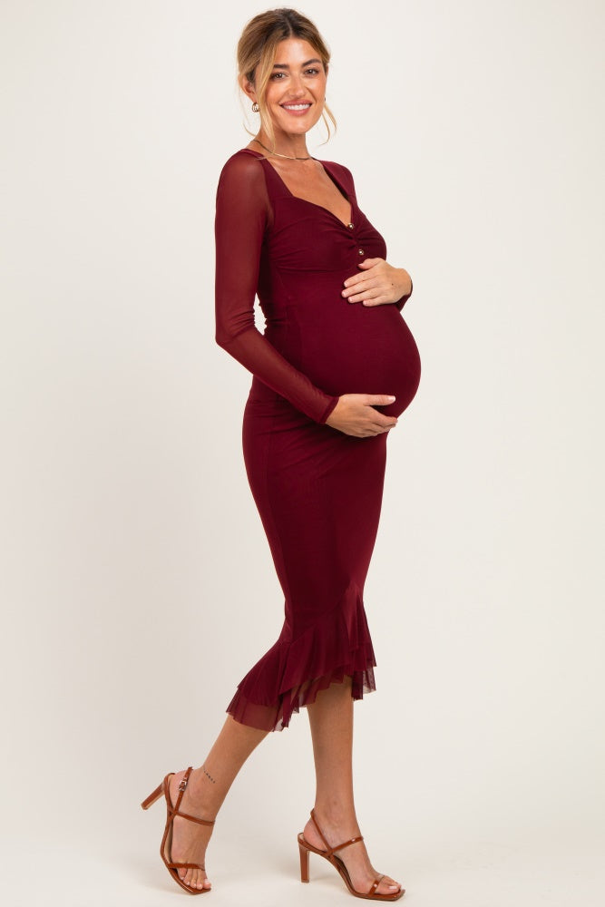 woman wearing a burgundy maternity dress with long sheer sleeves and a ruffled hem, holding her baby bump