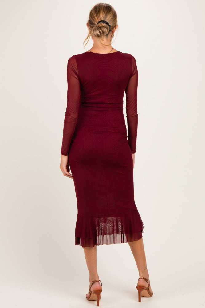 woman wearing a burgundy long sleeve maternity dress with a sheer ruffle hem and her back facing the camera