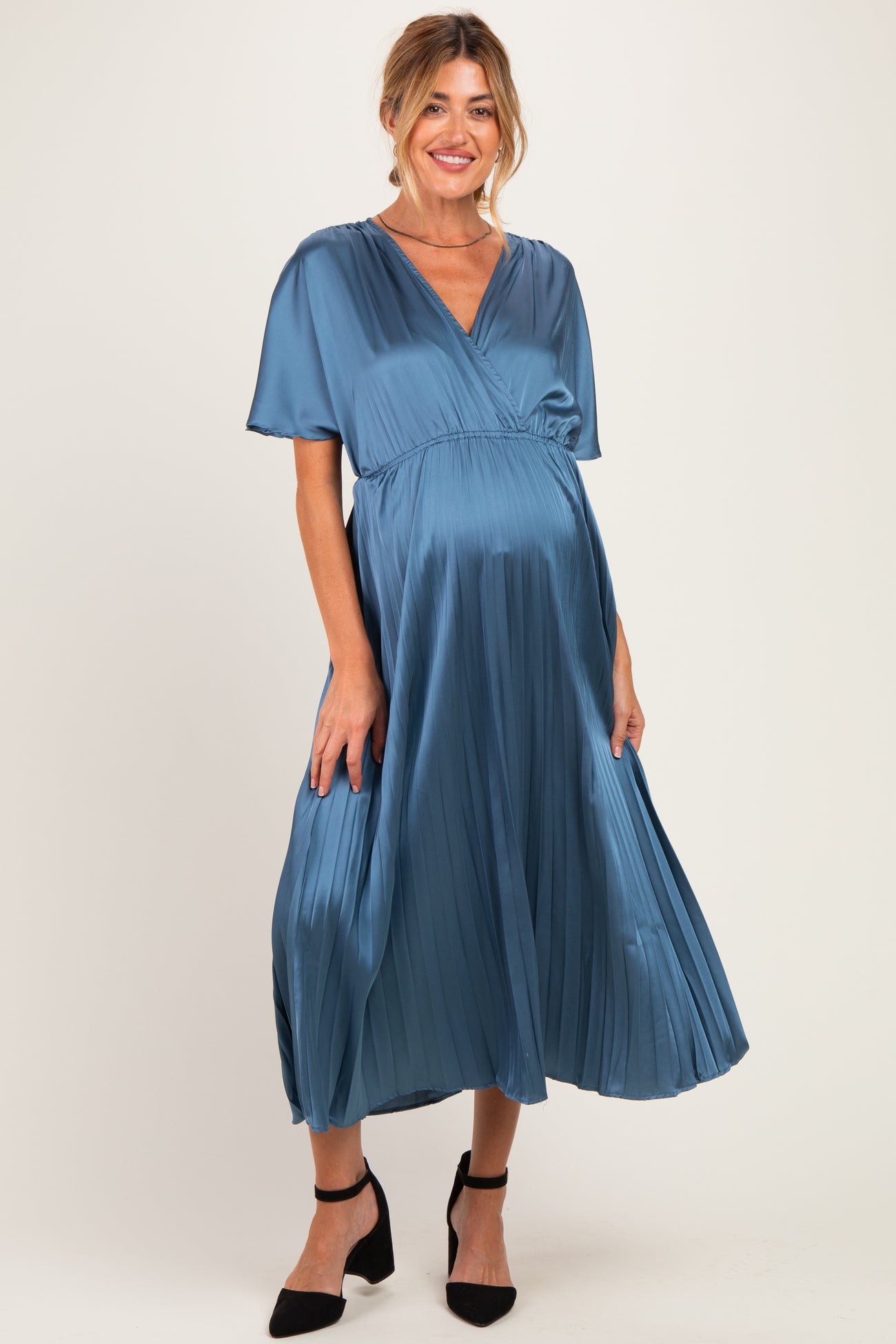 woman wearing a blue satin maternity dress with short sleeves and a pleated skirt, showcasing her baby bump