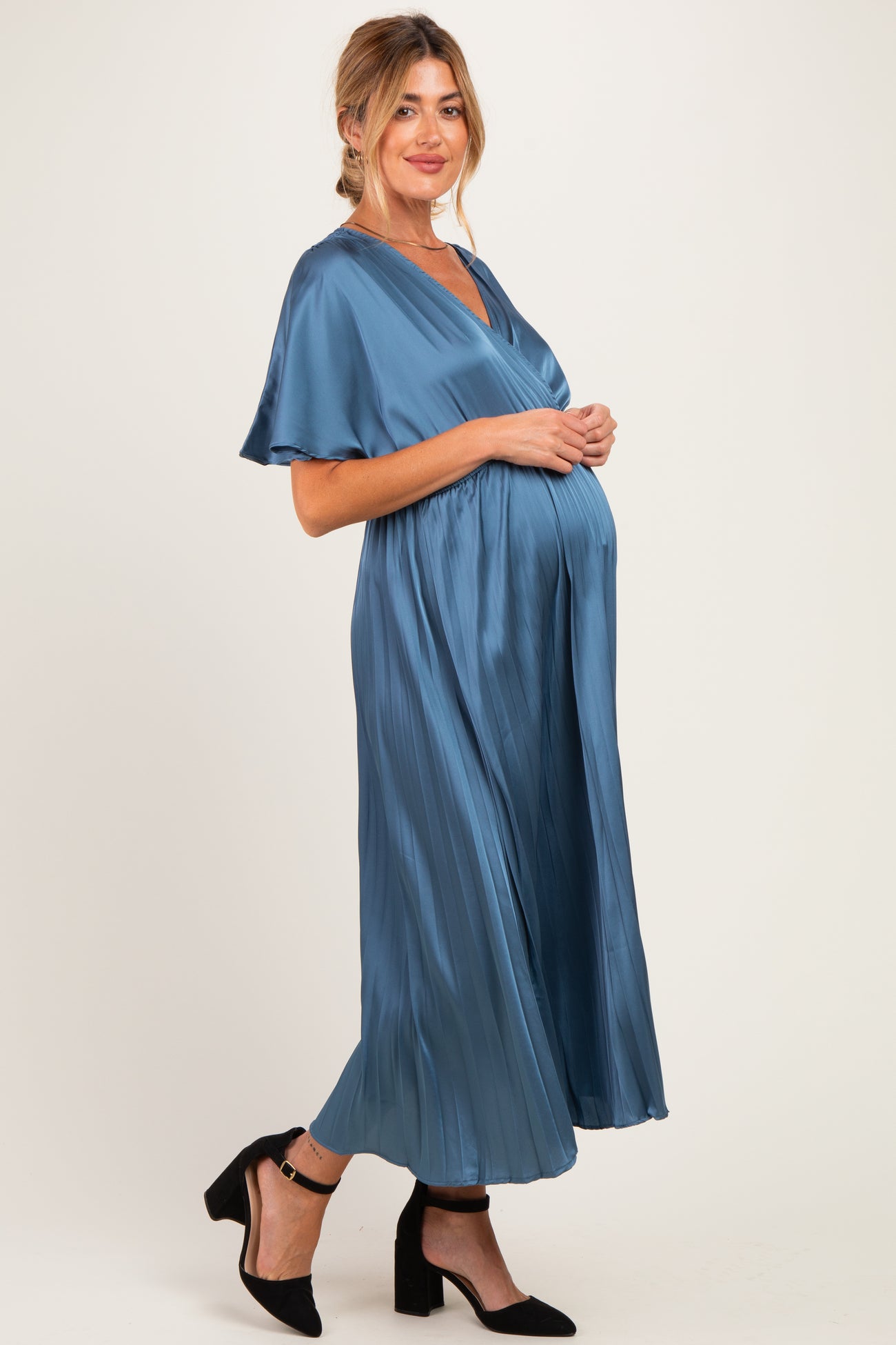 woman wearing a blue satin maternity dress with flutter sleeves and a flowing skirt, highlighting her baby bump