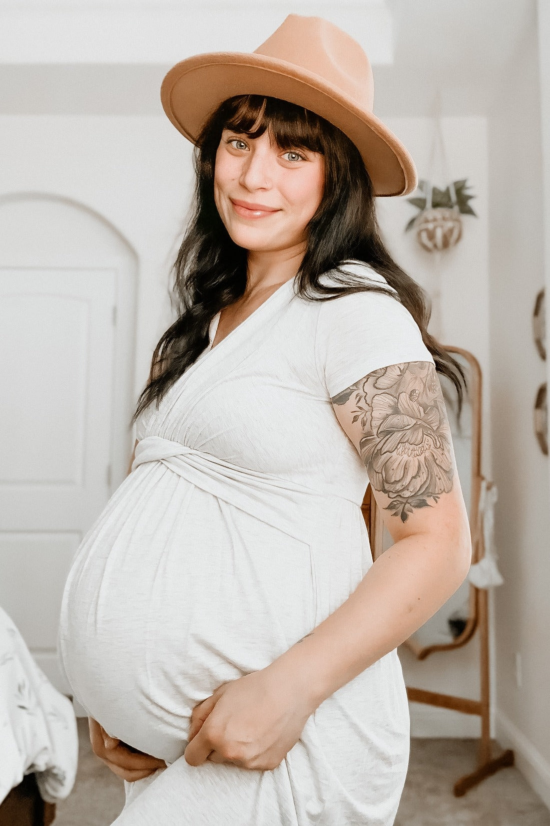 woman wearing a light gray maternity dress and a tan hat, showing her baby bump while smiling