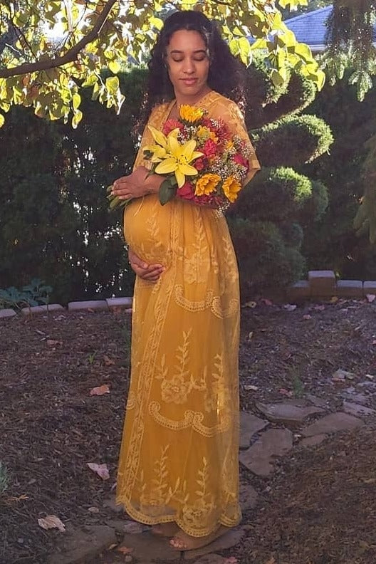 woman wearing a mustard yellow lace maternity dress, holding a bouquet and cradling her baby bump