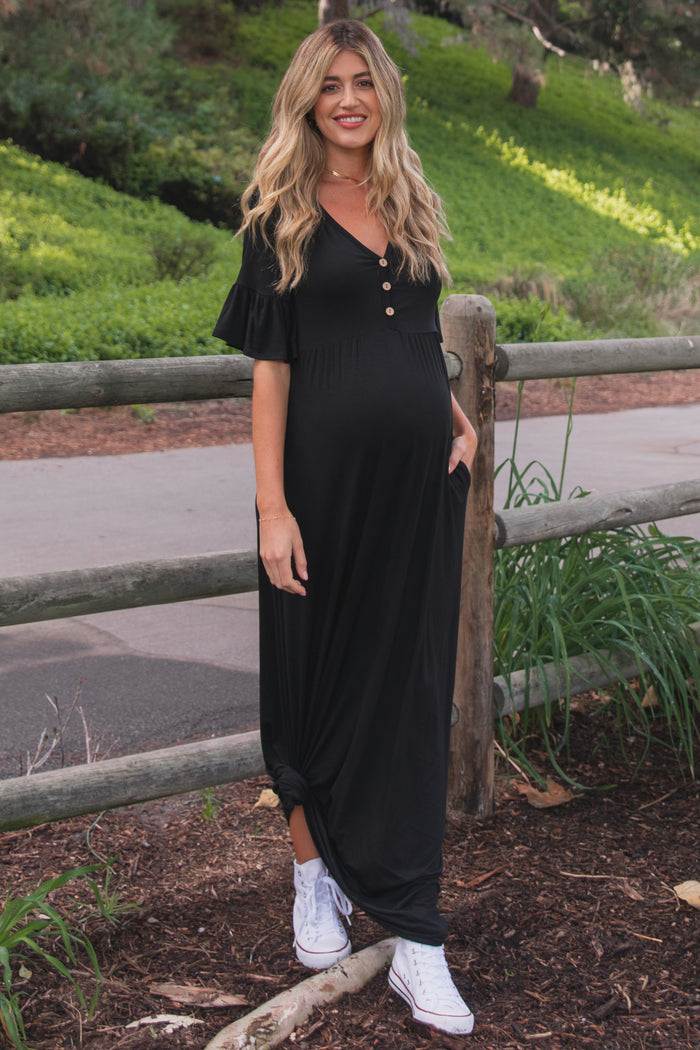 woman wearing a black maternity maxi dress with ruffled sleeves and buttons, showing her baby bump while standing outdoors