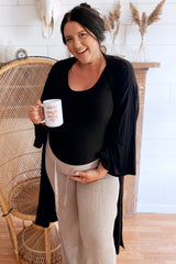 woman wearing a black maternity bodysuit and beige maternity pants, holding a mug and showing her baby bump